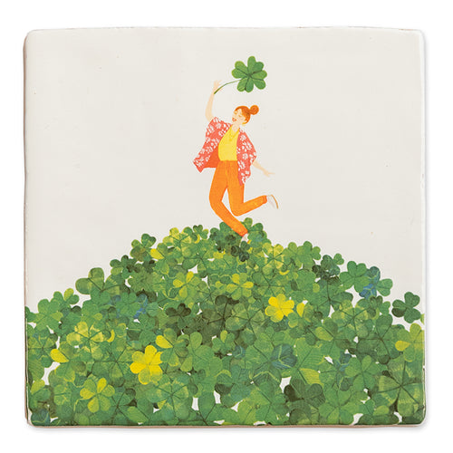 Storytile Vind je geluk. Mooie tegel met een vrouw in rood, geel en oranje kleding vrolijk dansend op een groen berg klavertjes vier. Ze heeft een groot klavertje vier in haar hand. 