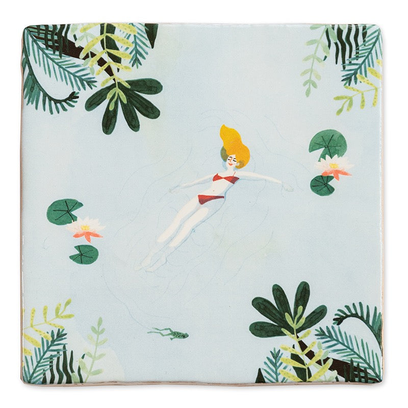 StoryTile Me-time. Op deze decoratietegel staat een vrouw die drijvend in het water lekker ontspant. Ze drijft tussen de waterlelies in een rode bikini. 
