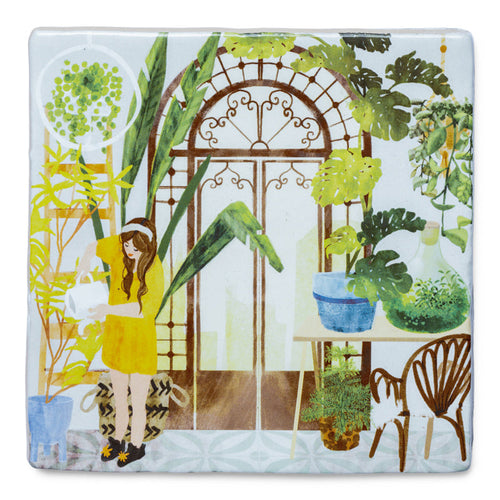 StoryTile Mijn groene oase. Ambachtelijk gebakken siertegel die een vrouw laat zien die in haar kas haar planten aan het verzorgen is. Ze draagt een gele jurk en is omringd met mooie planten. 